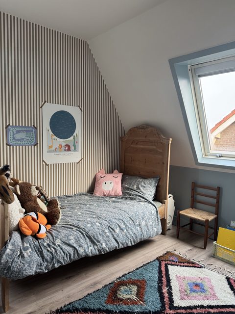 Kinderkamer met streepbehang, een oud houten bed, een rieten stoeltje en een Berber vloerkleed