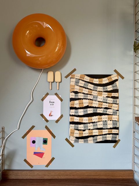 Oranje donutlamp aan een grijsblauwe muur, met tekeningen en een String-kast