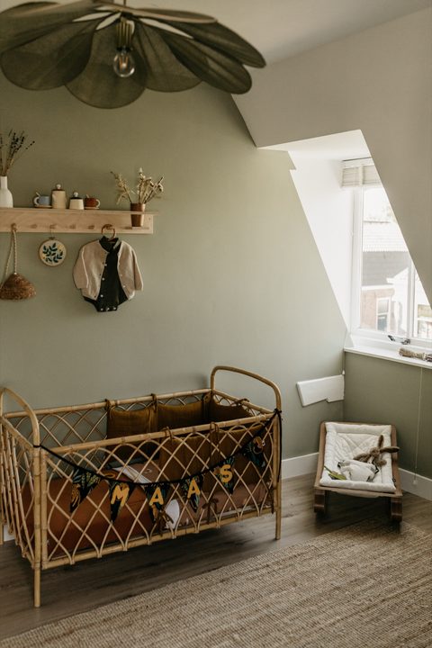 Babykamer in zachtgroen met een rotan ledikant, een houten wandplank en een slinger met de naam Maas