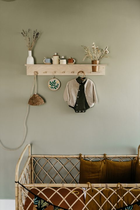 Houten wandplank met een vestje, droogbloemen en potjes boven een rotan ledikant