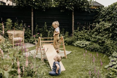 Twee kinderen spelen met een houten klimdriehoek in een groene tuin vol bloemen, met een rieten stoel